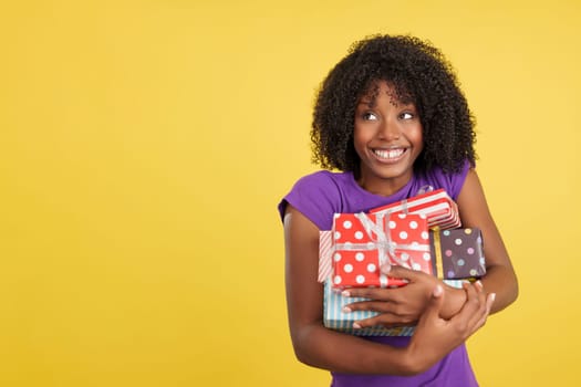 Funny and greedy woman with afro hair holding presents in studio with yellow background