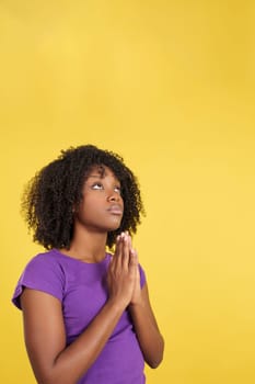 Afro woman praying while looking up joining hands in studio with yellow background