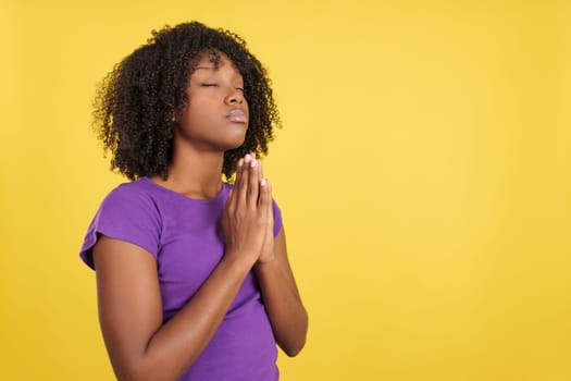 Woman with afro hair praying looking up with eyes closed and folded hands in studio with yellow background