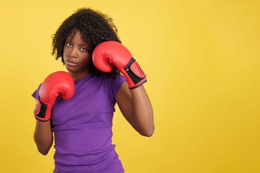 Woman with afro hairstyle posing with boxing gloves in studio with yellow background