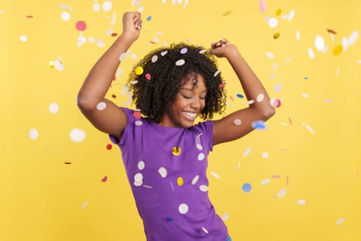 Cool Afro woman dancing and celebrating surrounded by confetti int he air in studio with yellow background
