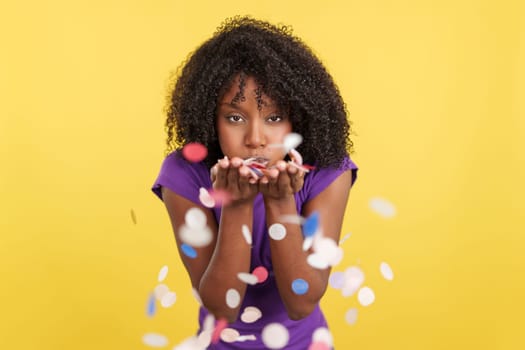 Cheerful Afro woman blowing confetti while celebrate in studio with yellow background