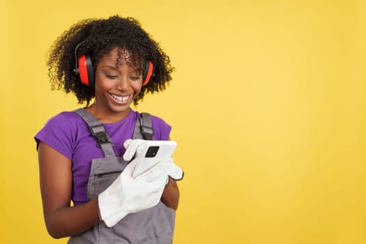 Afro female carpenter standing using the mobile in studio with yellow background