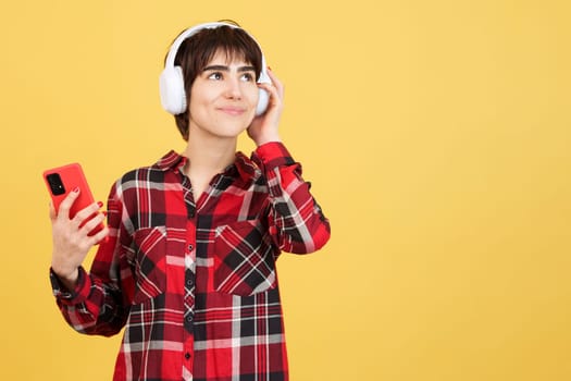 Happy androgynous person listening to music with mobile and headphones in studio with yellow background