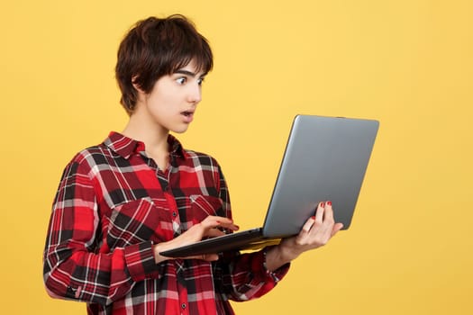 Surprised androgynous person standing and using a laptop in studio with yellow background