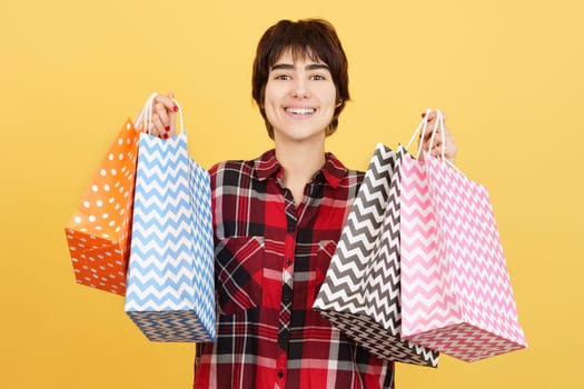 Happy androgynous person holding many shopping bags in studio with yellow background