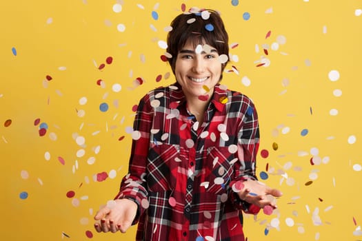 Happy androgynous person surrounded by flying confetti in studio with yellow background