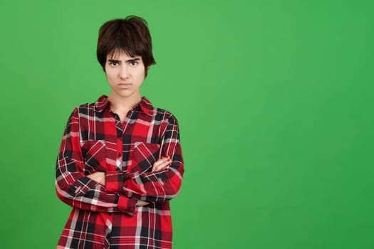 Upset androgynous person looking at the camera with angry expression and arms crossed in studio with green background