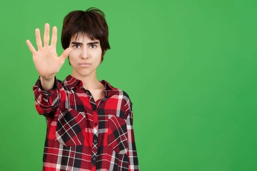 Serious androgynous person gesturing rejection with the palm of the hand in studio with green background