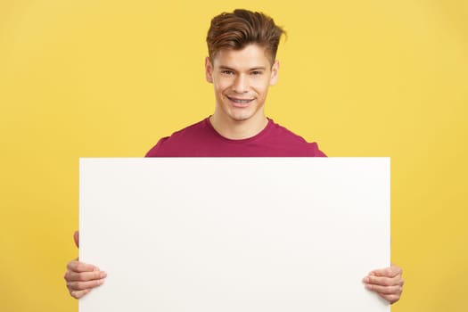 Caucasian man holding a blank panel smiling at the camera in studio with yellow background