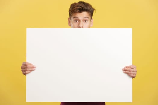 Surprised man hiding behind a blank panel in studio with yellow background