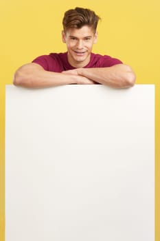 Blonde handsome man leaning on a blank panel in studio with yellow background