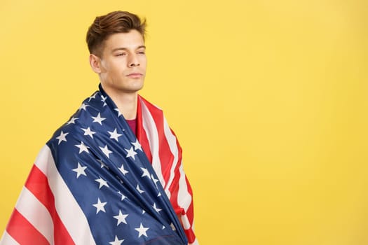 Handsome serious man wrapping with a United States national flag in studio with yellow background