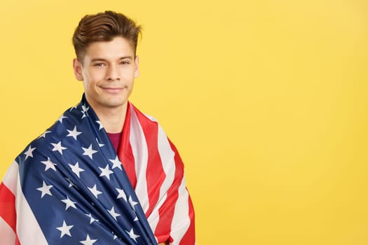 Proud man wrapping with a USA national flag in studio with yellow background