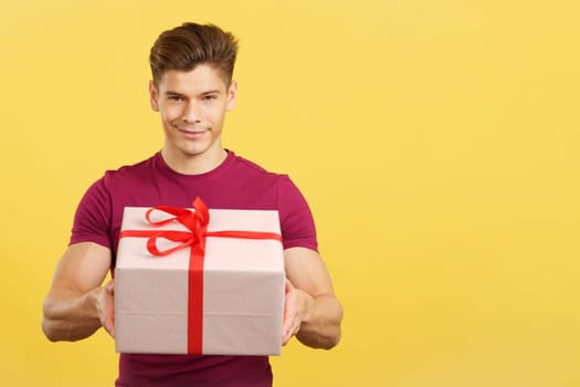 Happy man looking at the camera giving a present in studio with yellow background