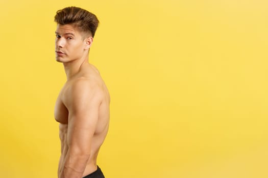 Handsome man with bare torso turning to looking at camera in studio with yellow background