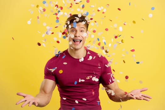 Cheerful man surrounded by confetti flying in the air in studio with yellow background