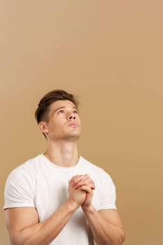 Handsome man looking up while praying with folded hands in studio with brown background