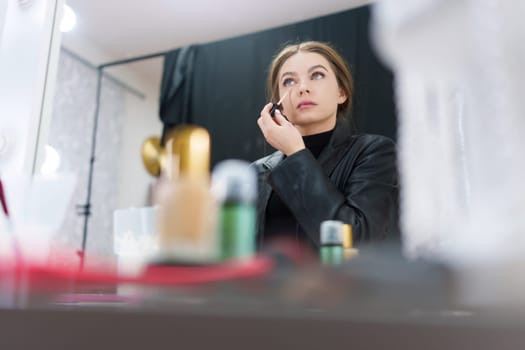 Low angle photo of a beauty woman applying concealer to under-eye circles sitting in front of a mirror in a backstage