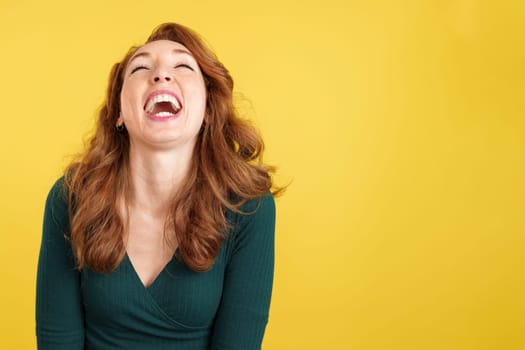 Happy redheaded woman laughing and looking up in studio with yellow background