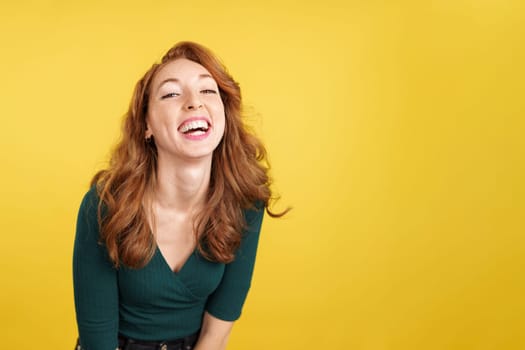 Happy redheaded woman laughing and looking at camera in studio with yellow background