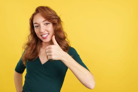 Happy redheaded woman gesturing agreement raising a thumb up in studio with yellow background