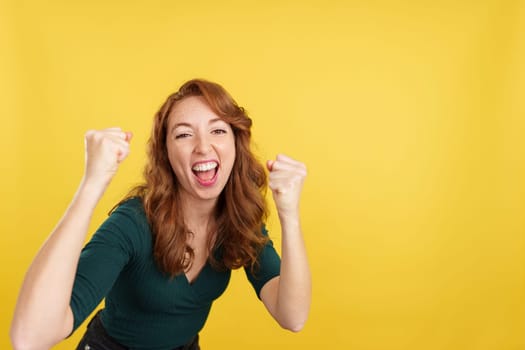 Happy redheaded woman celebrating raising the fists in studio with yellow background