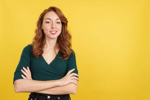 Redheaded woman standing with arms crossed smiling at the camera in studio with yellow background