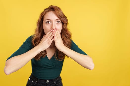 Redheaded woman keeping a secret while covering the mouth with hands in studio with yellow background
