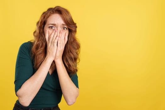 Redheaded woman covering the face in fear in studio with yellow background