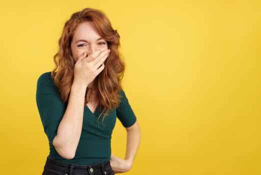 Shy redheaded woman covering the mouth while smiling in studio with yellow background