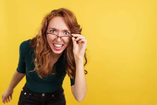 Redheaded woman with glasses looking at the camera with surprise in studio with yellow background