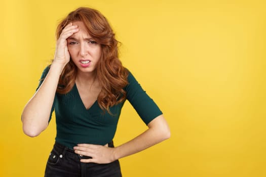 Redheaded woman gesturing bodily discomfort and headache in studio with yellow background