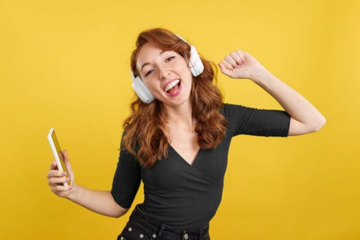 Redheaded happy woman listening to music and dancing using headphones and a mobile in studio with yellow background