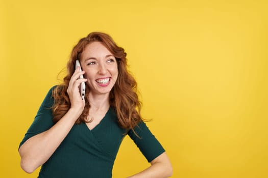 Happy redheaded woman talking to the mobile in studio with yellow background