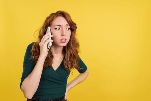 Worried redheaded woman talking to the mobile in studio with yellow background