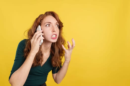 Angry redheaded woman talking to the mobile in studio with yellow background