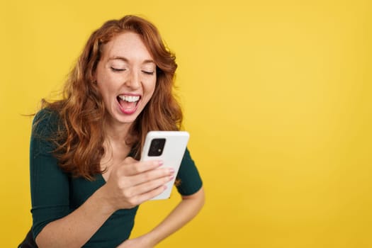 Happy Redheaded woman laughing while using the mobile phone in studio with yellow background