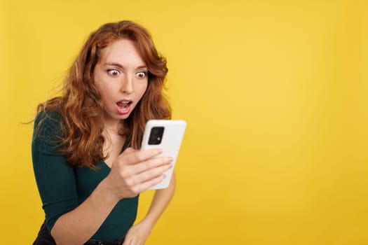 Surprised redheaded woman looking the screen of a mobile in studio with yellow background