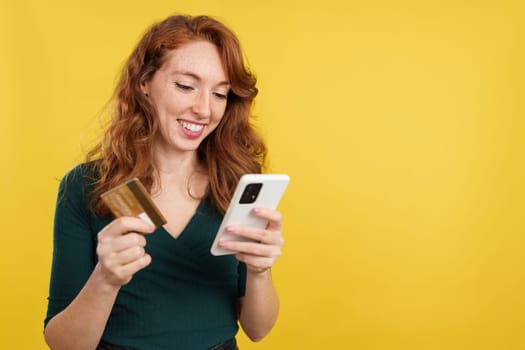 Happy redheaded woman shopping online with the mobile and a card in studio with yellow background