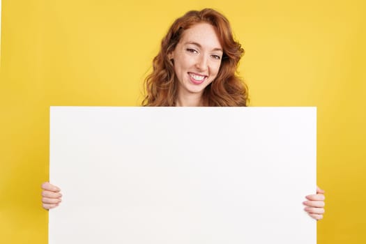 Redheaded woman holding a blank panel while smiling at the camera in studio with yellow background