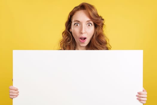 Amazed redheaded woman holding a blank panel while looking at camera in studio with yellow background