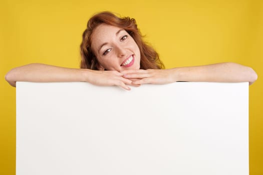 Relaxed redheaded woman leaning on a blank panel in studio with yellow background