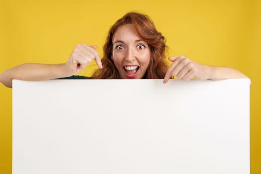 Amazed redheaded woman pointing to a panel with blank space in studio with yellow background
