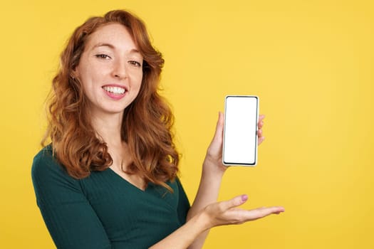 Redheaded happy woman presenting a mobile with a blank screen in studio with yellow background