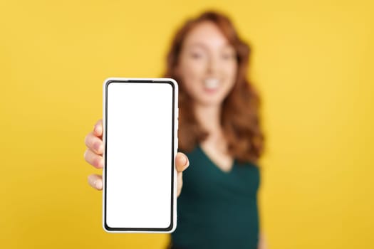 Blank screen of the mobile held by a redheaded woman in studio with yellow background