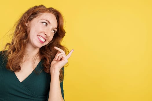 Happy redheaded woman pointing a blank space to the side in studio with yellow background