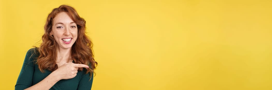 Redheaded woman pointing a blank space and smiling in studio with yellow background