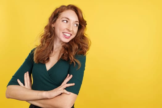 Redheaded woman standing with arms crossed smiling in studio with yellow background