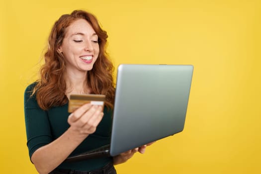 Redheaded woman shopping online with a laptop and a credit card in studio with yellow background